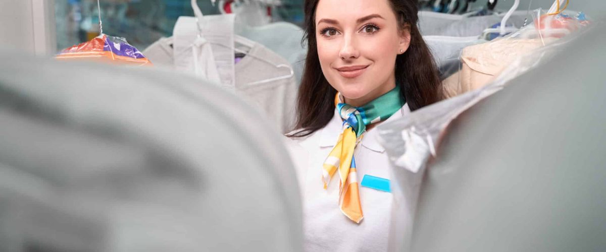 Female administrator of laundry looking for needed items walking along racks with garment on hangers, professional dry cleaning service