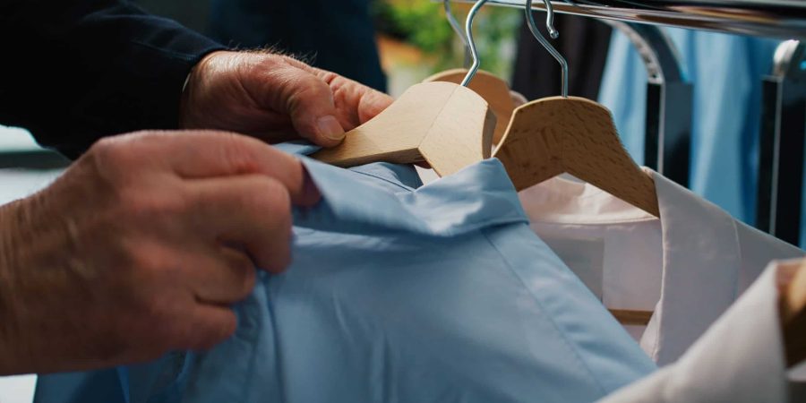 Elderly client checking all clothes placed on display at showroom, browsing through hangers to find items on discount. Senior man shopping for elegant shirts and suit jackets. Close up. Camera B.